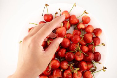 Çekim, canlı kırmızı kiraz yığınının ortasında, tombul, taze bir kiraz için uzanan bir eli yakalar, basit bir zevki ve sağlıklı beslenmeyi hatırlatır. Yemek blogları, sağlıklı tarifler veya meyve promosyonları için ideal.