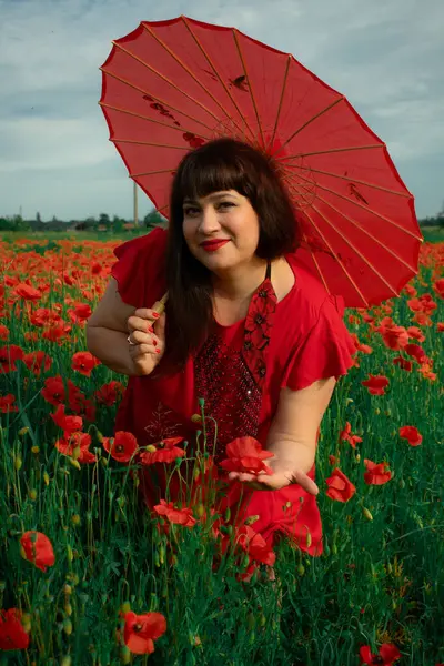 Kırmızı elbiseli ve şemsiyeli bir kadın canlı bir çiçek tarlasında haşhaşı tutarken büyüleyici bir şekilde gülümsüyor. Yaz temaları, güzellik ve güzel doğa deneyimleri için ideal.