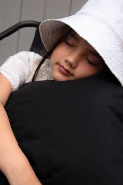 Şapkalı genç bir kız huzurlu bir şekilde kestiriyor, sakin bir ortamda yumuşak siyah bir yastığa sarılıyor. Çocukluk temaları, rahatlama, sakin anlar, aile, rüyalar veya huzur verici görseller için mükemmel