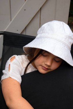 Şapkalı genç bir kız huzurlu bir şekilde kestiriyor, sakin bir ortamda yumuşak siyah bir yastığa sarılıyor. Çocukluk temaları, rahatlama, sakin anlar, aile, rüyalar veya huzur verici görseller için mükemmel