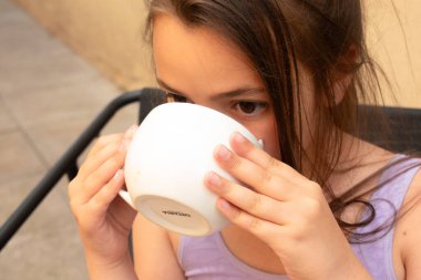 Güneşli bir bahçede büyük beyaz bir bardaktan su içen bir kız. Sakin bir yaz sabahı, çocuklar için sıcaklığı ve sağlıklı yaşam tarzını sembolize ediyor. Çocuk sofra takımı markaları için yaz kampanyaları