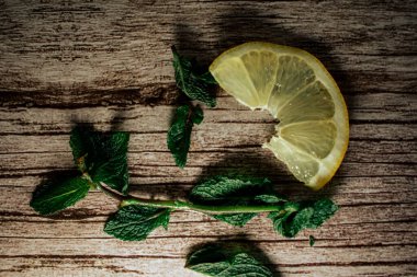 Taze limon dilimleri ve canlı nane filizleri kırsal bir ahşap yüzeye saçılmıştır. Bu kompozisyon doğal tazeliği ve sağlıklı malzemeleri çağrıştırır, içecek, yiyecek veya yaz temaları için mükemmeldir.