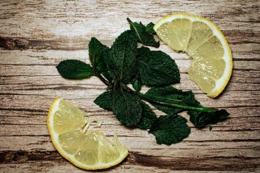 Taze limon dilimleri ve canlı nane filizleri kırsal bir ahşap yüzeye saçılmıştır. Bu kompozisyon doğal tazeliği ve sağlıklı malzemeleri çağrıştırır, içecek, yiyecek veya yaz temaları için mükemmeldir.