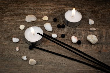 Düz taşlar ve siyah tütsü çubuklarıyla çevrili, ahşap bir yüzeyde parlak bir mum yanar. Rahatlama, farkındalık ve iç huzuru çağrıştıran doğal bir sahne..