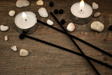 Düz taşlar ve siyah tütsü çubuklarıyla çevrili, ahşap bir yüzeyde parlak bir mum yanar. Rahatlama, farkındalık ve iç huzuru çağrıştıran doğal bir sahne..