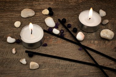 Düz taşlar ve siyah tütsü çubuklarıyla çevrili, ahşap bir yüzeyde parlak bir mum yanar. Rahatlama, farkındalık ve iç huzuru çağrıştıran doğal bir sahne..