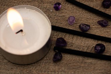 Düz taşlar ve siyah tütsü çubuklarıyla çevrili, ahşap bir yüzeyde parlak bir mum yanar. Rahatlama, farkındalık ve iç huzuru çağrıştıran doğal bir sahne..