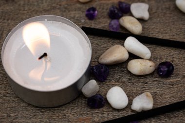 Düz taşlar ve siyah tütsü çubuklarıyla çevrili, ahşap bir yüzeyde parlak bir mum yanar. Rahatlama, farkındalık ve iç huzuru çağrıştıran doğal bir sahne..