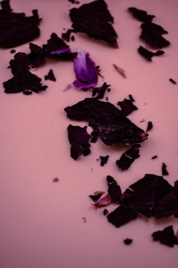 Kurumuş koyu yapraklar ve yaprak parçaları açık pembe bir yüzeye dağılmış, parlak mor bir taç yaprağı ise kontrastı ekliyor. Yumuşak, organik zarafeti olan doğal, dokusal bir kompozisyon.