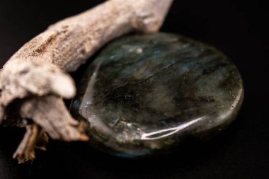 Mavi-yeşil labradorit taş koyu arkaplandaki desenli dalların yanında duruyor. Parlak kristal ve kırsal ahşap kontrastı refah ve dekor temaları için doğal bir kompozisyon oluşturur.