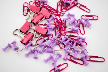 Kırmızı ataçlar ve mor raptiyeler beyaz arkaplana saçılmış, kısmen görünür pembe bir ataç ile. Ofis, okul ve organizasyon temaları için renkli bir kırtasiye karışımı