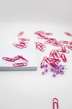 Kırmızı ataçlar ve mor raptiyeler beyaz bir yüzeye saçılmış boş bir kağıt yığınının yanında. Organizasyon ve yaratıcılık kavramları için ofis malzemelerinin renkli ve dinamik düzenlenmesi