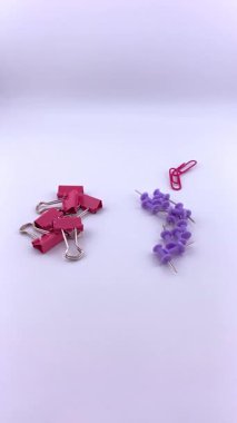 Kırmızı ataçlar ve mor raptiyeler beyaz arkaplana saçılmış, kısmen görünür pembe bir ataç ile. Ofis, okul ve organizasyon temaları için renkli bir kırtasiye karışımı