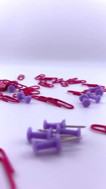 Kırmızı ataçlar ve mor raptiyeler beyaz arkaplana saçılmış, kısmen görünür pembe bir ataç ile. Ofis, okul ve organizasyon temaları için renkli bir kırtasiye karışımı