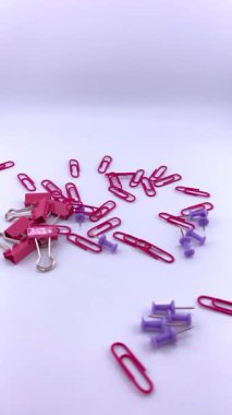 Kırmızı ataçlar ve mor raptiyeler beyaz arkaplana saçılmış, kısmen görünür pembe bir ataç ile. Ofis, okul ve organizasyon temaları için renkli bir kırtasiye karışımı