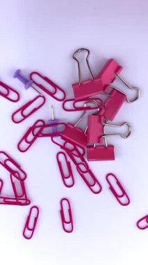 Kırmızı ataçlar ve mor raptiyeler beyaz arkaplana saçılmış, kısmen görünür pembe bir ataç ile. Ofis, okul ve organizasyon temaları için renkli bir kırtasiye karışımı