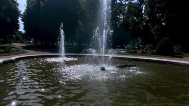 Serinletici fıskiye jeti mavi gökyüzüne ve yeşil ağaçlara karşı yükseliyor, güneş ışığında dalgalanmalar ve su yüzeyine sıçramalarla parıldıyor. Yaz doğası arka planı.