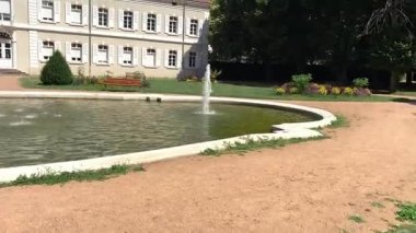 İki katlı geniş bir binanın geniş açılı manzarası güneş panelleri, fayanslı bir çatı ve güneşli bir gölete bakan beyaz panjurlar. Huzurlu bir ortamda çeşmeler, banklar ve çalılar..