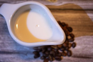 Kremalı beyaz seramik krema ve ahşap üzerine serpiştirilmiş kahve çekirdekleri. Sıcak dokular ve yumuşak ışık kahve ve yaşam tarzı temaları için mükemmel rahat, sade bir atmosfer yaratır.