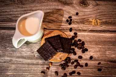 Dekoratif bantlı beyaz seramik sürahi, ahşap dilimli koyu çikolata ve kırsal ahşap üzerine yayılmış kahve çekirdekleri. Sıcak, samimi ve davetkar bir yaşam tarzı..