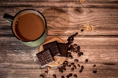 Etrafı dağınık fasulyelerle çevrili, ahşap bir bardak altlığın üzerinde koyu çikolata kareleri olan bir fincan kahve. Kırsal ahşap ve sıcak ışık hoş, davetkar bir atmosfer yaratır..