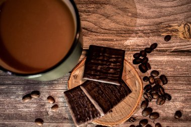 Etrafı dağınık fasulyelerle çevrili, ahşap bir bardak altlığın üzerinde koyu çikolata kareleri olan bir fincan kahve. Kırsal ahşap ve sıcak ışık hoş, davetkar bir atmosfer yaratır..