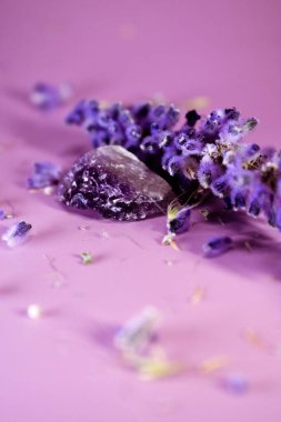 Pembe yüzeyde etrafa saçılmış lavanta çiçekleri olan Ametist kristalleri. Yansıtıcı arkaplan mor renkleri, doğal güzelliği, dinginliği ve ruhani uyumu harmanlıyor..
