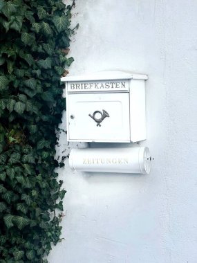 BRIEFKASTEN yazılı beyaz bir posta kutusu ve beyaz duvarında ZEITUNGEN yazan bir gazete konteynırı. Klasik bir kornanın içinde bir mücevher kutusu, yakınlarda yeşil sarmaşık yetişiyor.,