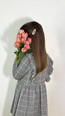 Fotoğrafta, uzun saçlı ve tokalı, kareli elbiseli bir kız görülüyor. Elinde bir buket pembe lale tutuyor. Arkaplan beyaz.
