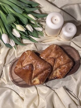 Açık saten bir kumaşa yerleştirilmiş, tahta bir tahtanın üzerinde üst üste dizilmiş iki kek. Yakınlarda bir buket beyaz lale ve iki dekoratif mum var..