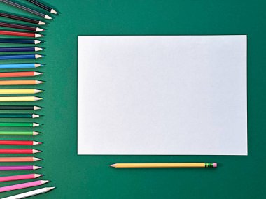 Yeşil bir yüzeye boş bir kağıt yerleştirilir. Etrafında çeşitli renkli kalemler var, yaratıcı öğrenme ve sanatsal ifade için hazırlar. Yüksek kalite fotoğraf