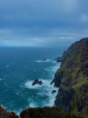 İrlanda Atlantik kayalıkları ve kıyı şeridi ile çarpan dalgalar, yeşil alanlar ve vahşi Atlantik yolu boyunca fırtınalı gökyüzü.