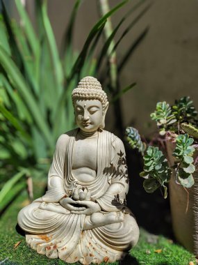 Serin yeşil bitkilerin arasında duran güzel bir Buda heykeli. Yumuşak ışık, gölgeler heykel üzerinde dans ederken yüz detaylarını ön plana çıkarıyor. Meditasyon için huzurlu ve uyumlu bir alan yaratıyor..