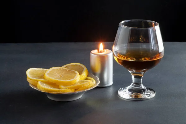 There is a glass of cognac on the table. Nearby are sliced lemons in a plate. A candle burns at the back.