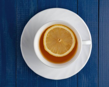 Masada bir fincan çay var. İçinde bir dilim limon yüzüyor. Yukarıdan bak. Mavi bir arkaplanda.