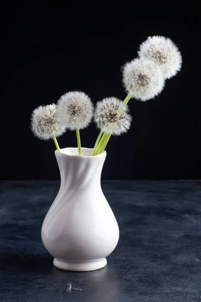 Masada beyaz bir vazoda karahindibalar var. Siyah bir arkaplanda.