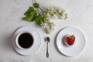 Masanın üzerinde bir fincan kahve, çilekli bir kek ve beyaz bir çiçek var. Beyaz bir arkaplanda.