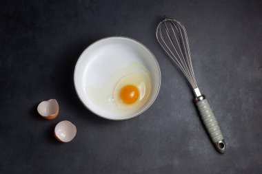 Masanın üzerinde kırık yumurtalı bir tava var. Yakınlarda bir yumurta kabuğu ve çırpıcı var. Siyah bir arkaplanda.