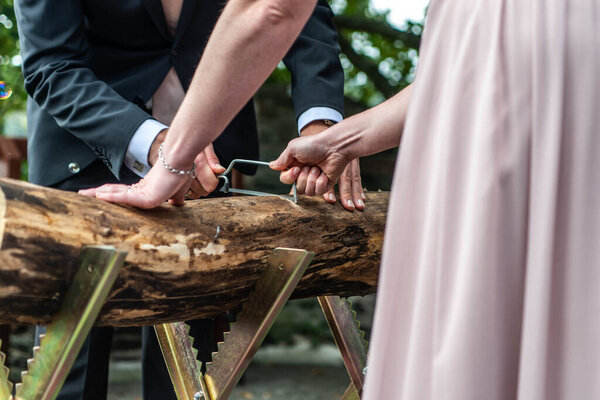 Young bridal couple groom bride sawing a tree trunk together german wedding tradition.
