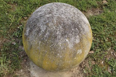 Çimenli bir zeminde sıkıca duran, yosun ve renk değişimleriyle yıpranmış beton bir kürenin yakın çekimi. Bu küresel boğa, sert, eski yüzeyi ve organik dokusuyla zamanın geçişini sergiliyor.. 