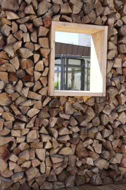 Ortasında ahşap çerçeveli kare bir pencere ile düzgünce düzenlenmiş bir odun yığını. Pencere, arka planda modern bir cam yapıyı ortaya çıkararak, kırsal doğal dokular ile kentsel mimari arasında sanatsal bir tezat oluşturuyor.. 