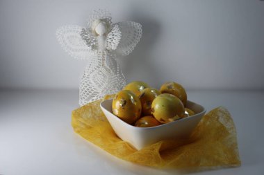 Bu dingin yaşam, yumuşak sarı bir kumaşa nazikçe yerleştirilmiş altın folyo aksanlı bir kase altın sarısı Paskalya yumurtası yakalıyor. Arka planda, beyaz el işi bir melek. 