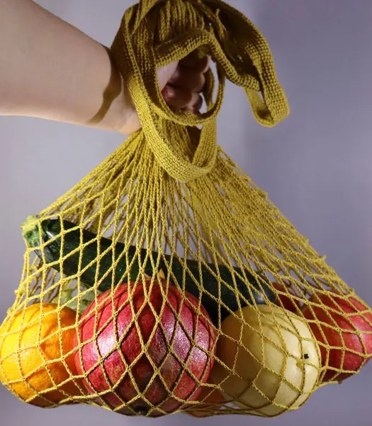 Bir el, taze meyve ve sebzelerle dolu, nar, portakal, elma ve kabak gibi yeniden kullanılabilir sarı bir ağ torbasını nötr bir zemin üzerinde tutar..
