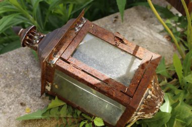 Yeşil bitkilerle çevrili eski paslı bir fener. Çürümeyi, klasik cazibeyi ve açık havada geçen zamanı simgeliyor..