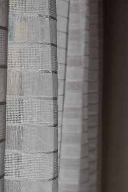 Doğal ışıkla hafifçe aydınlatılan hassas ızgara desenli zarif perdelerin yakın çekimi. Minimalist iç tasarım elementi sakin bir atmosfer yaratıyor.