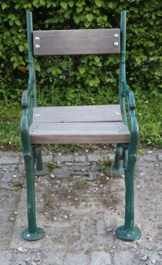 Yeşil bir parktaki taş kaldırımda duran tek bir ahşap ve metal sandalye. Klasik açık hava koltukları doğal gün ışığında çimenler ve çalılarla çevrilidir..