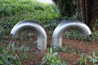 Yeşil bitkiler ve ağaçlarla çevrili bir bahçede yerden çıkan iki kıvrımlı metal boru. Modern endüstriyel tasarım doğal ortamda.