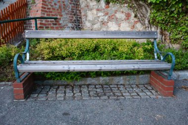 Metal kollukları ve tuğla destekleri olan basit bir ahşap bank sarmaşıkla kaplı ve yeşil çalılarla çevrili taş bir duvarın yanına yerleştirilmiş..