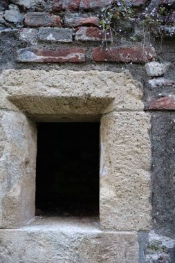 Tarihi tuğla bir duvarda eski bir taş pencere çerçevesi. Küçük dikdörtgen açıklık yıpranmış taşlar ve eski tuğlalarla çevrili. Bu da kırsal ve ortaçağ atmosferi yaratıyor..