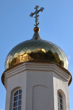 Beyaz bir kilise kulesinin tepesinde açık mavi gökyüzüne karşı kurulmuş Hıristiyan Ortodoks haçı olan altın bir soğan kubbesinin yakın görüntüsü. İnancın ve mimarinin sembolü..